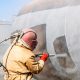 Un travailleur industriel en tenue de protection utilisant une sableuse pour nettoyer et préparer une surface métallique dans un atelier.